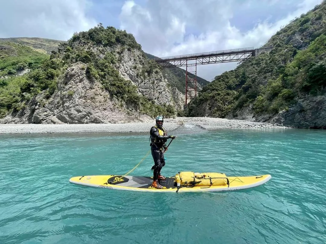 Tony Pain co-founder in wetsuit and helmet paddleboarding on turquoise river with rocky and green hills, and a bridge in the background.
