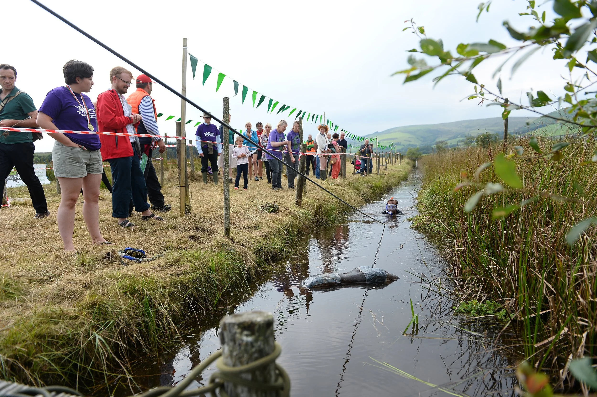 A group of people standing along the edge of a narrow waterway, with one person in the water wearing a snorkel and mask. The scene is outdoors, with green hills in the background and green flags suspended above the group.