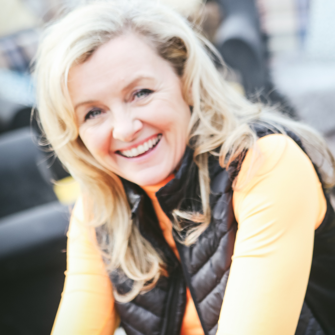 Smiling woman with blonde hair in a black vest and orange long-sleeve shirt, indoors.