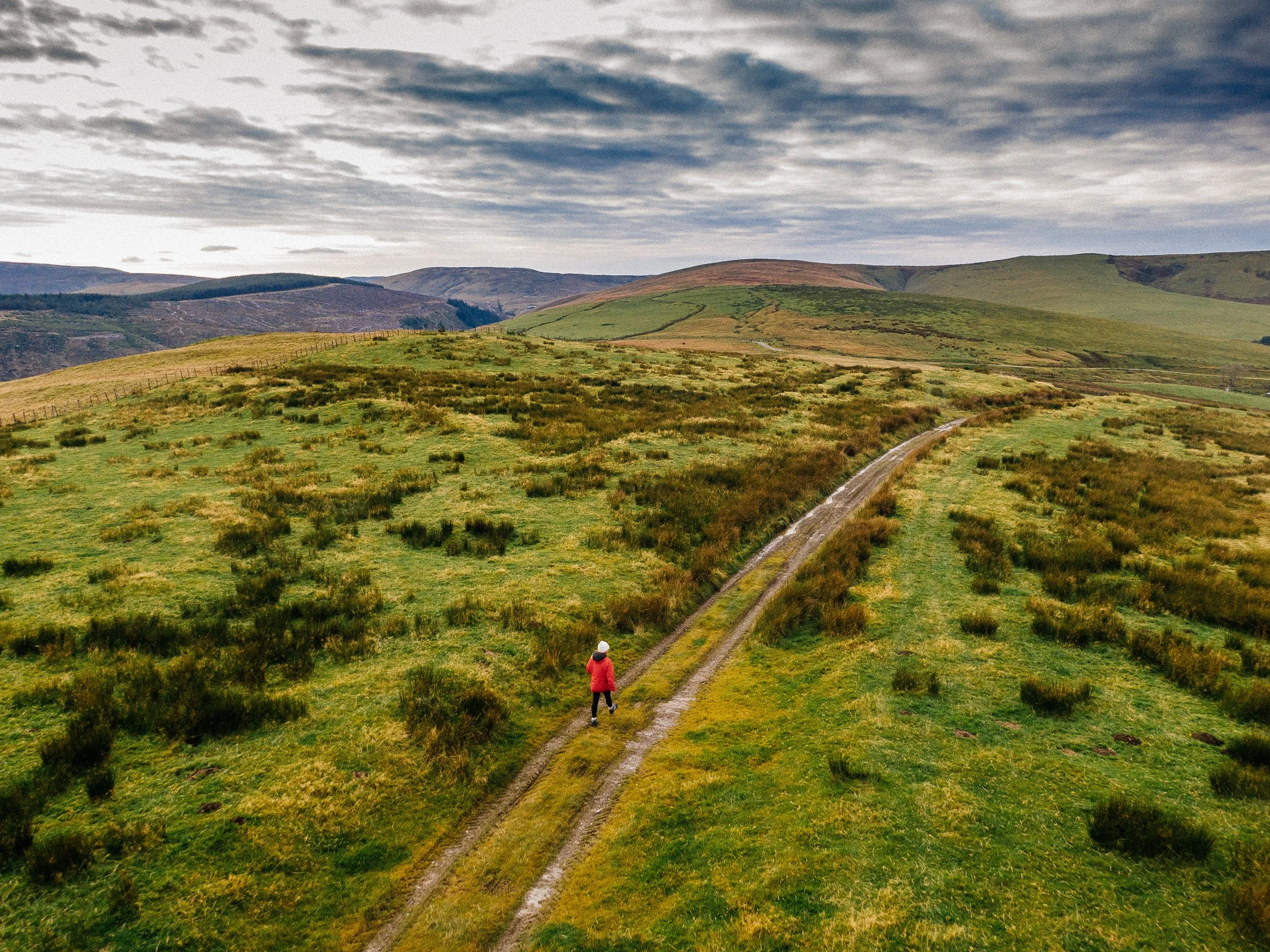 A person in a red jacket walking up a dirt path in a grassy hillside with rolling green hills and cloudy sky in the background.