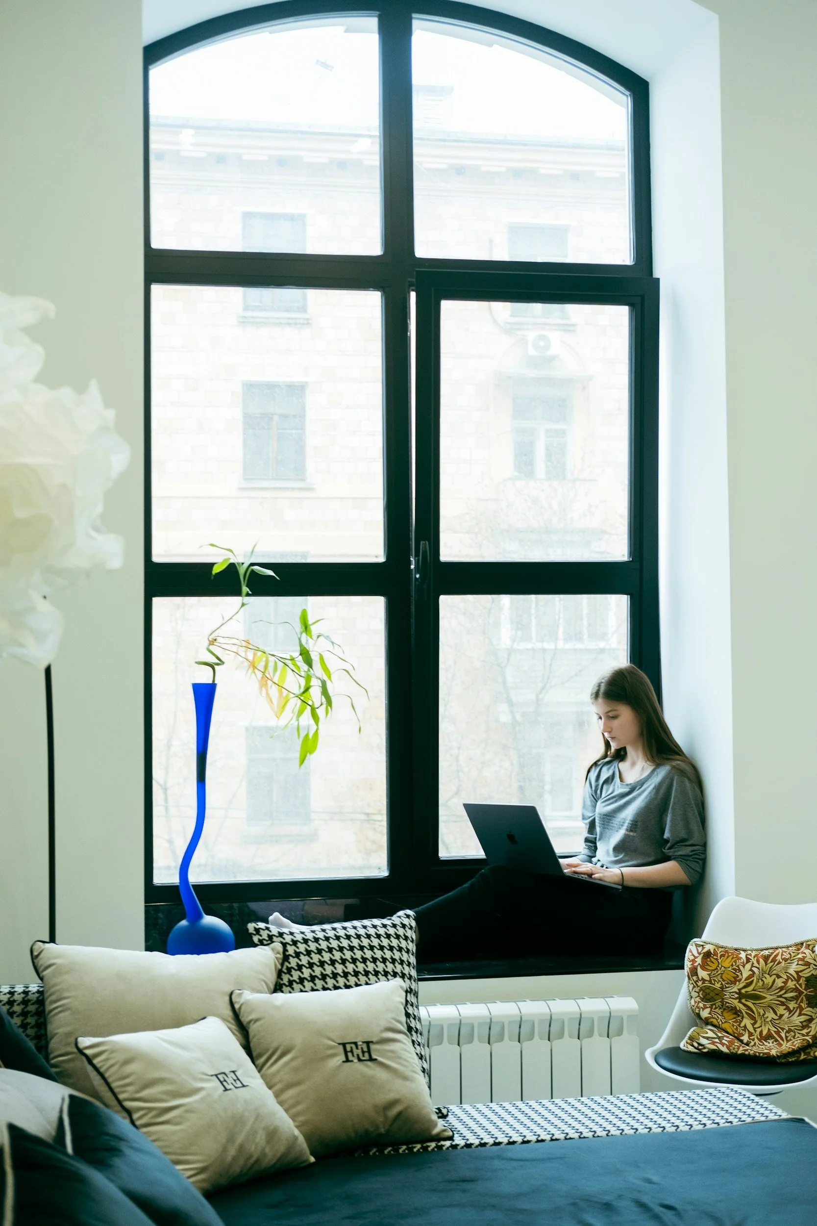 A woman sitting on a window seat working on a laptop in a bright room with large arched windows, a blue vase with a plant on the windowsill, and home decor including pillows and a chair with a colorful cushion.