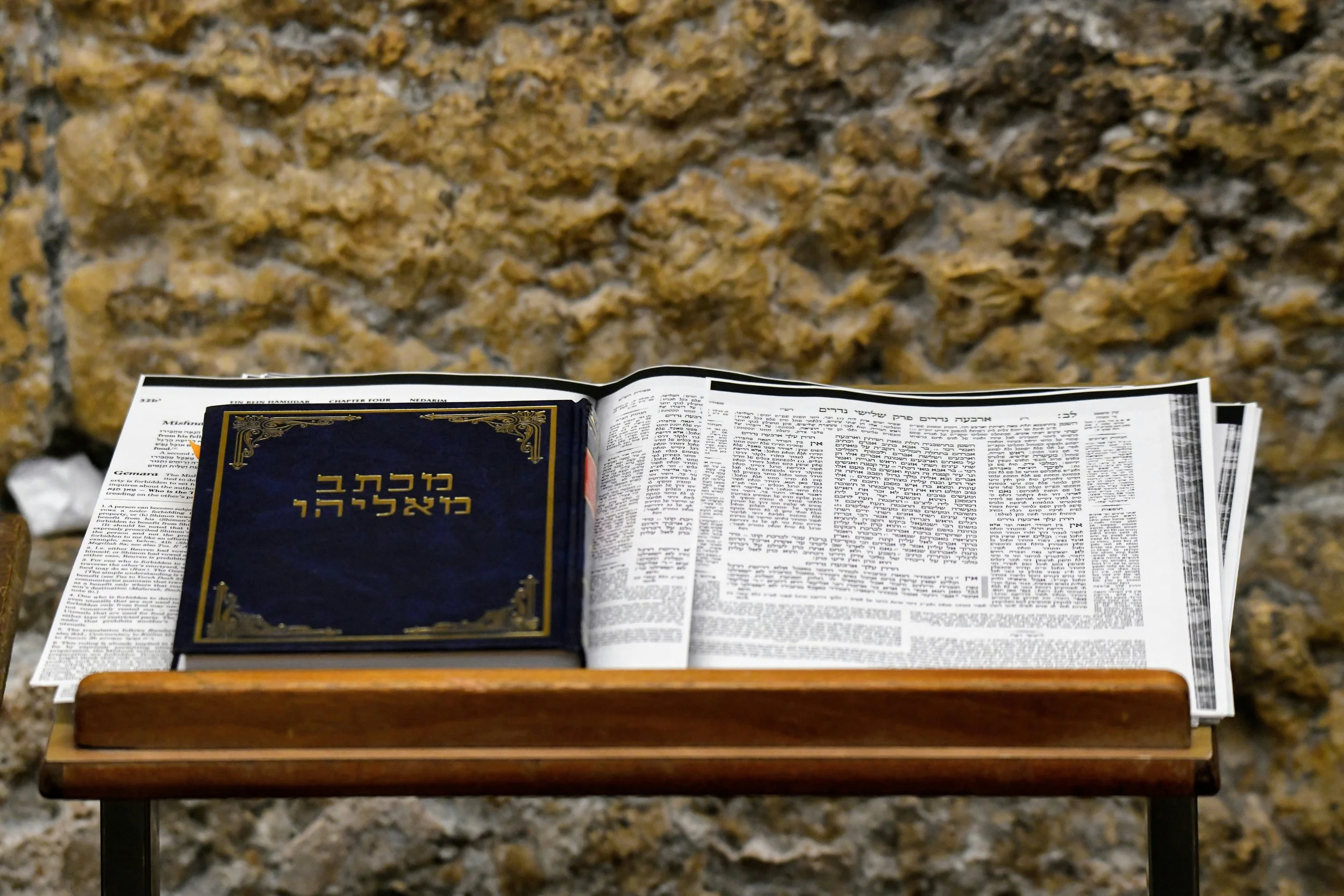 Open book with Hebrew text on a wooden stand, placed on a table with a stone wall in the background.