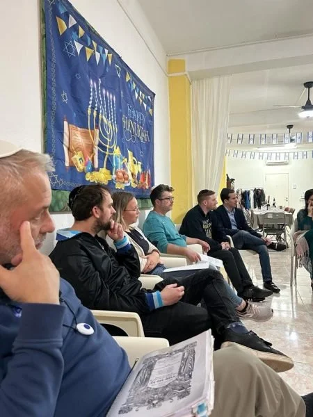 People sitting in a row in a room decorated for Hanukkah with a blue banner, menorah, and Hanukkah symbols on the wall.