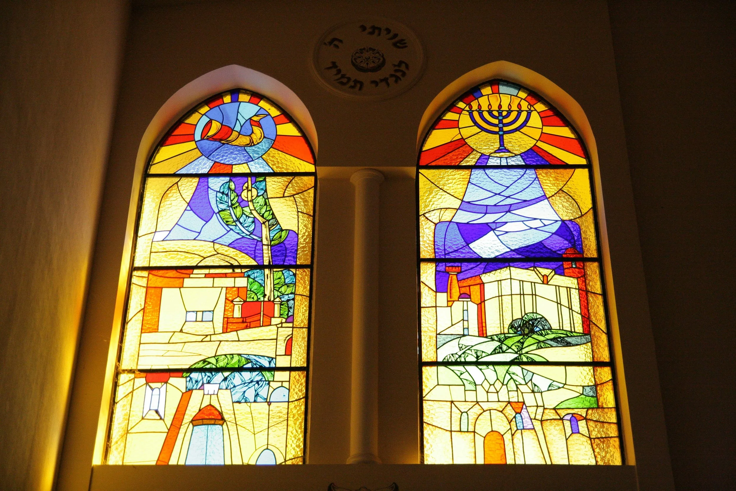 Colorful stained glass windows depicting various religious symbols and scenes, including a menorah and a shofar, in a religious setting.