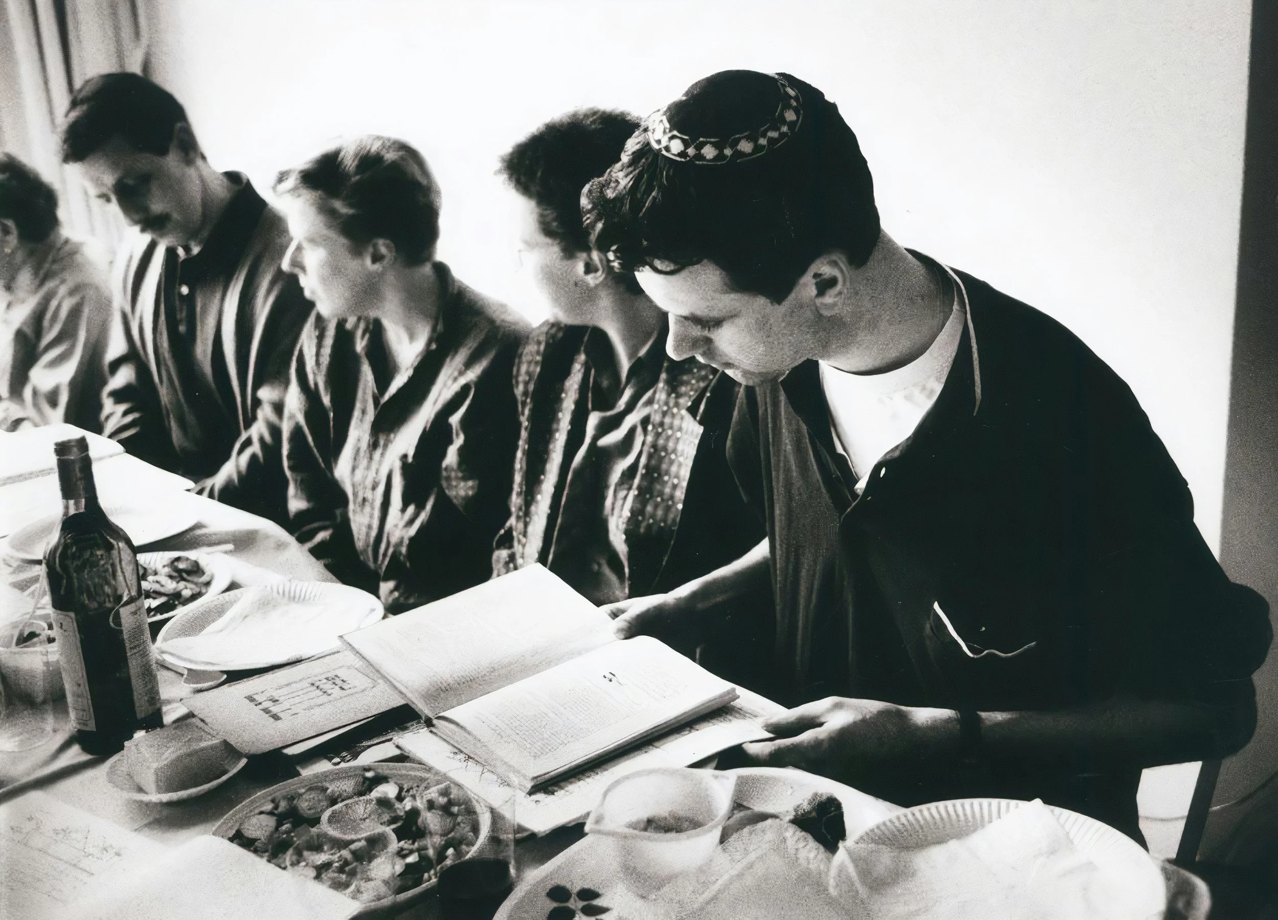 People sitting at a table with food and drinks, appearing to participate in a meal or gathering, in a black and white photo.