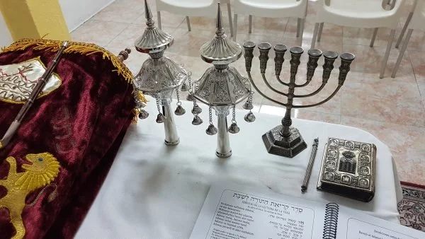 Jewish ceremonial items on a table, including two silver Torah crowns, a menorah, a prayer book, and a notebook.