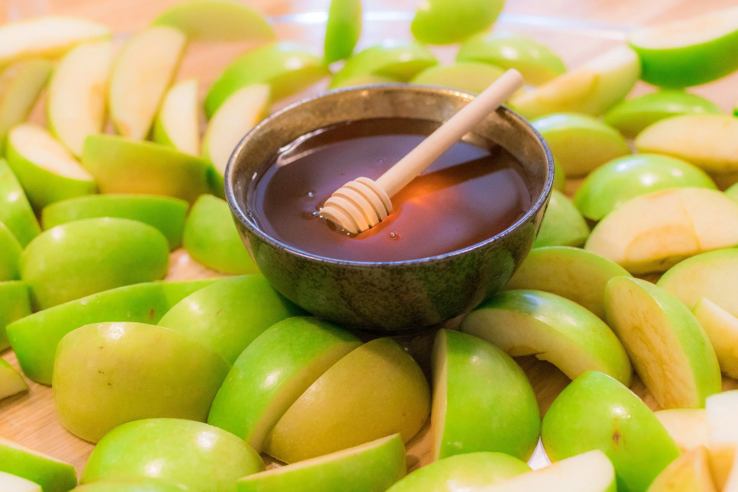 A bowl of honey with a honey dipper resting inside, surrounded by sliced green apples on a wooden surface.
