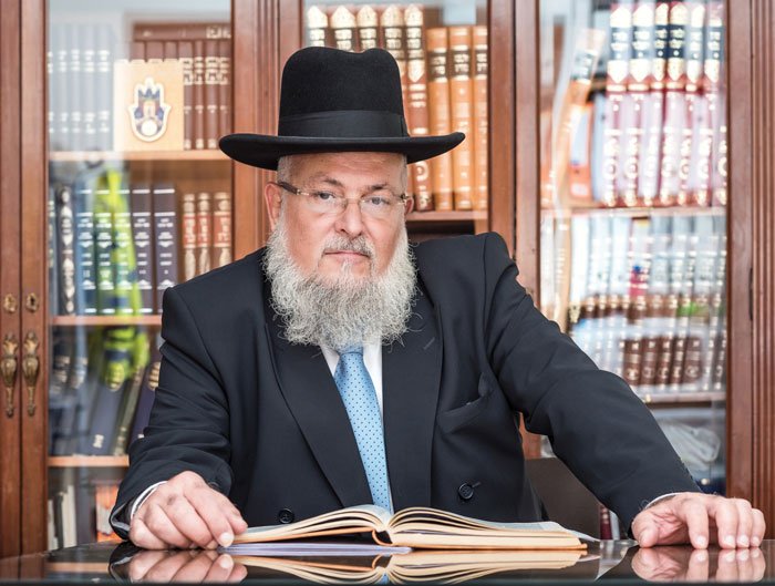 A man wearing a black hat and suit sitting at a desk in a library, with an open book in front of him.