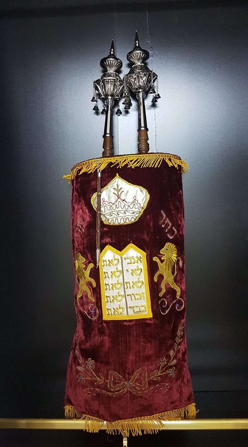 A Torah scroll adorned with a red velvet cover embroidered with gold designs, flanked by two silver finials, and attached to a wooden staff. The cover features Hebrew inscriptions and decorative lions.