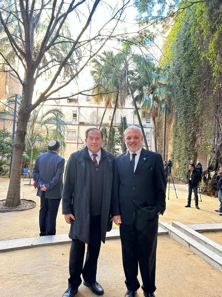 Two men in suits standing outdoors in a garden, with other people and camera equipment in the background, among trees and plants.