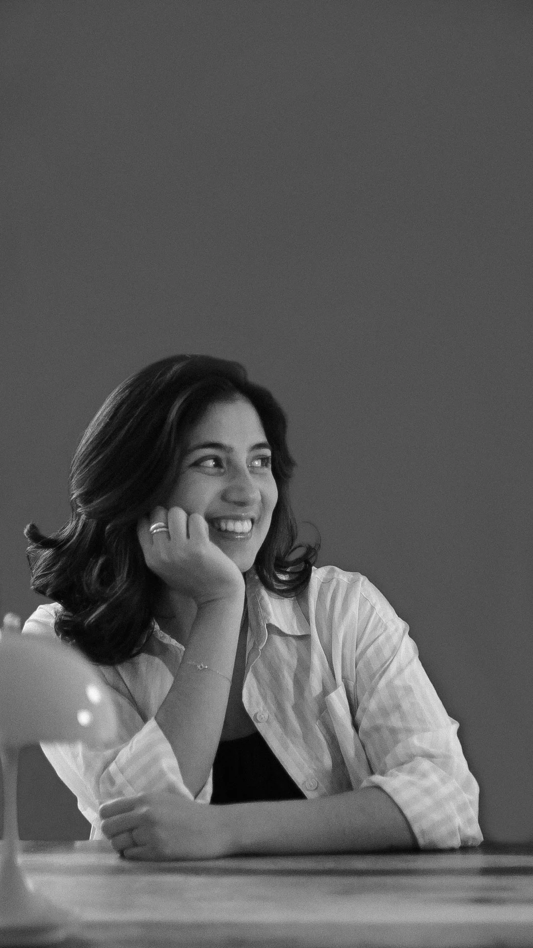 Black and white photo of a smiling woman with dark wavy hair, resting her chin on her hand, wearing a light-colored shirt, sitting at a table.