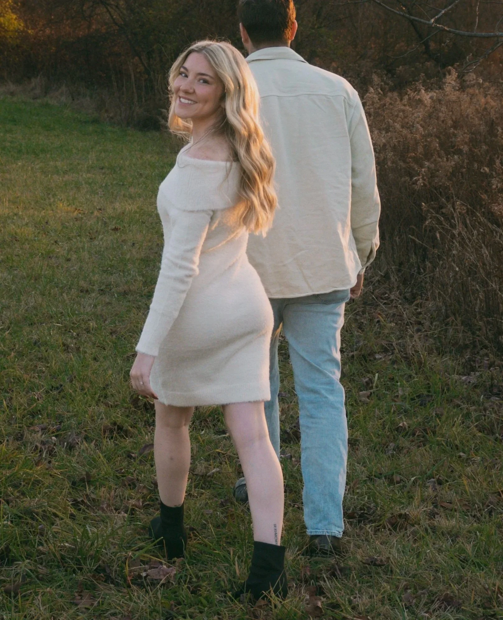A woman with long blonde hair smiling and looking back at the camera, standing in a grassy field during sunset, with a man facing away from the camera next to her, in a natural outdoor setting.
