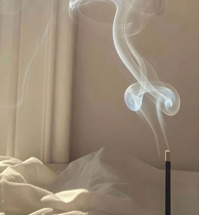 A lit incense stick releasing swirling smoke in a room with cream-colored walls and a bed with white sheets.
