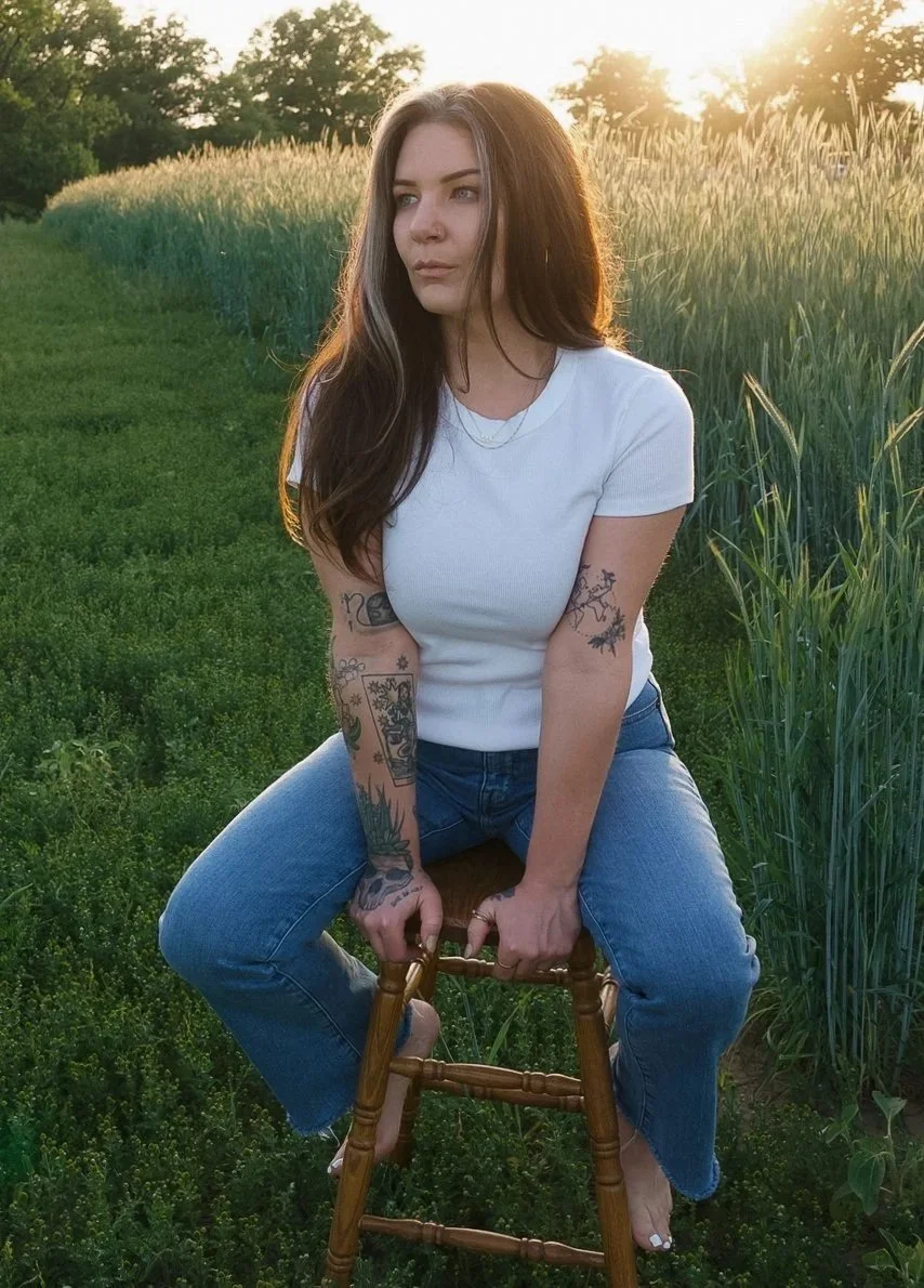 A young woman with long brown hair and tattoos on her arms, wearing a white t-shirt and jeans, sitting on a wooden stool in a grassy field during sunset.