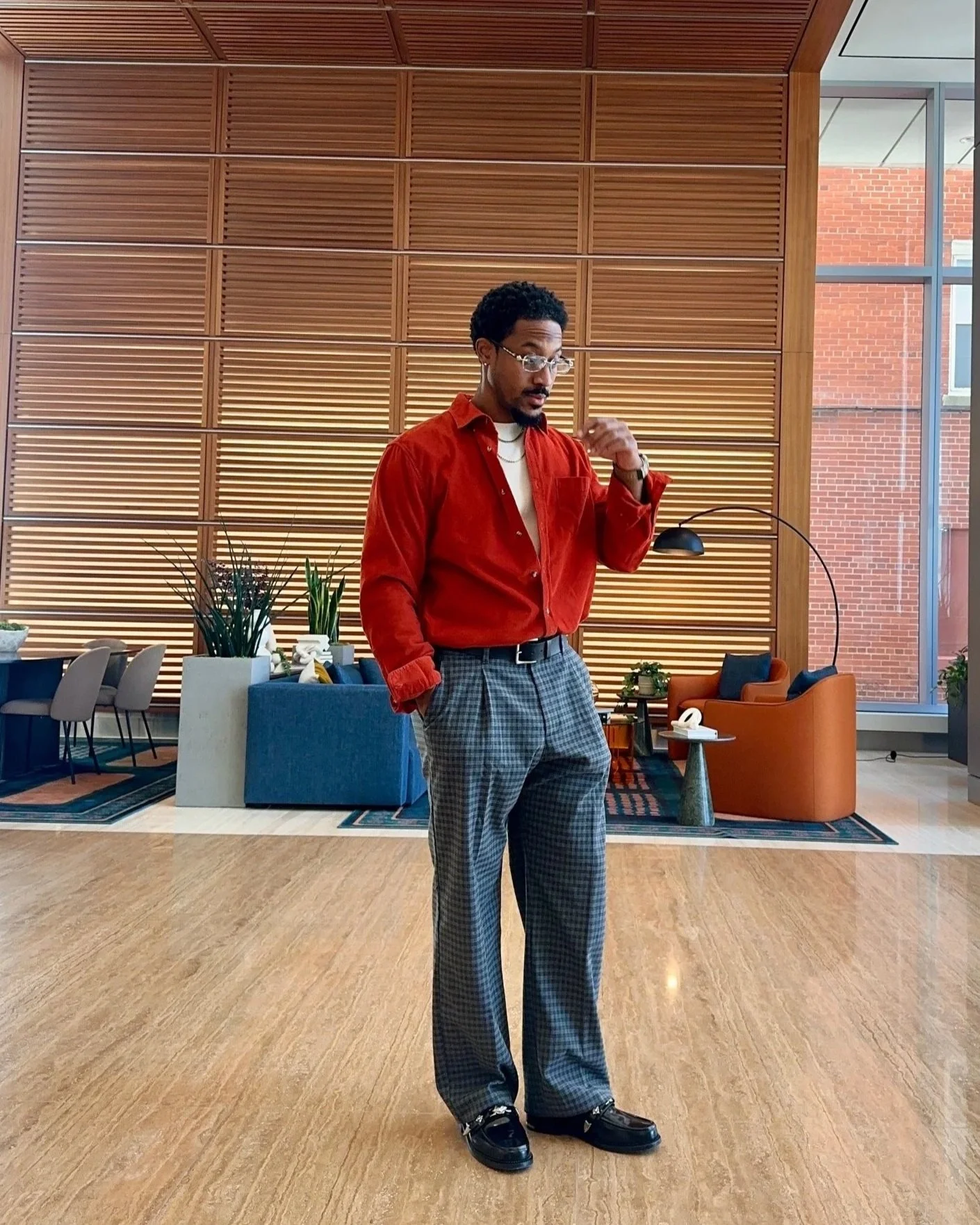 A man with glasses and curly hair wearing a red shirt, checkered brown and black pants, and black shoes standing in a modern interior space with wooden wall panels, plants, and seating area.