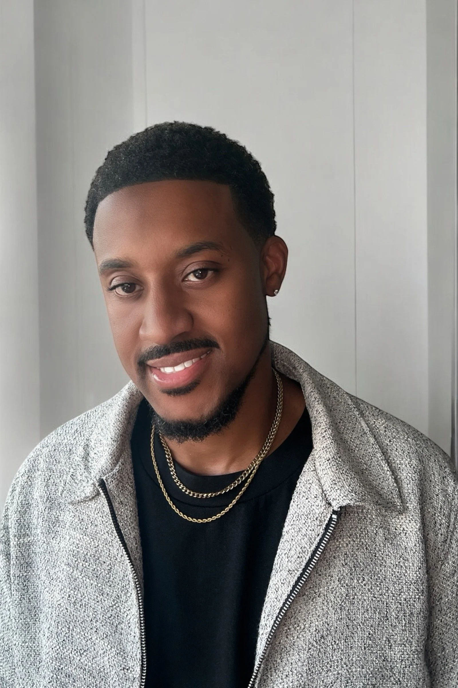 A young man with black hair, a mustache, and a goatee, smiling, wearing a gray textured zip-up jacket over a black shirt, with layered gold chains around his neck, standing in front of a white wall.
