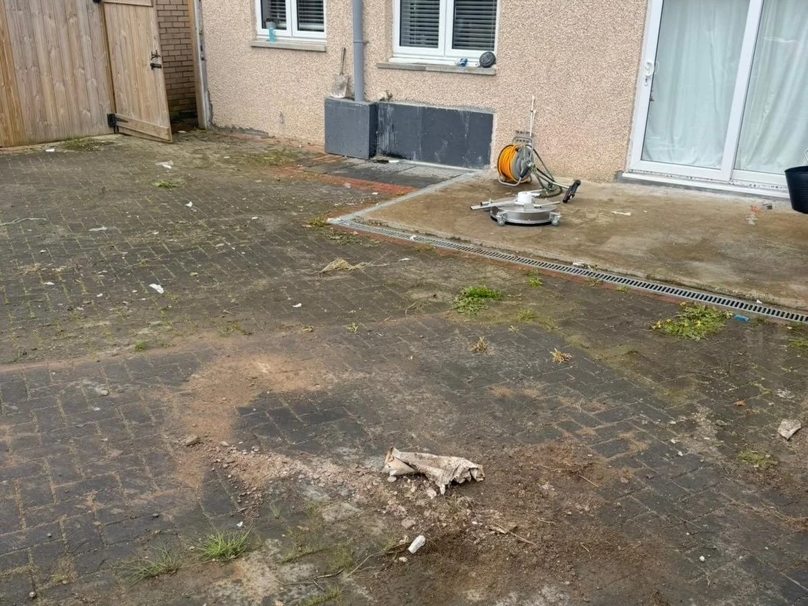Backyard with paving stones, some areas under construction or renovation, with construction tools and equipment nearby, and a small patch of grass and weeds.