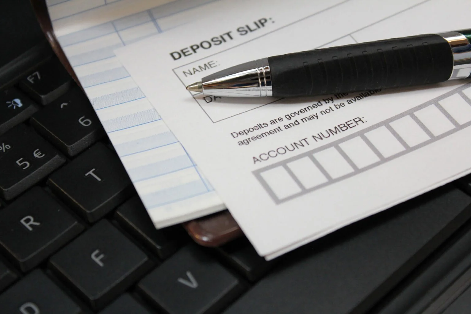 A deposit slip with a black and silver pen rests on a computer keyboard, partially covering a paper with spaces for name, date, and account number.