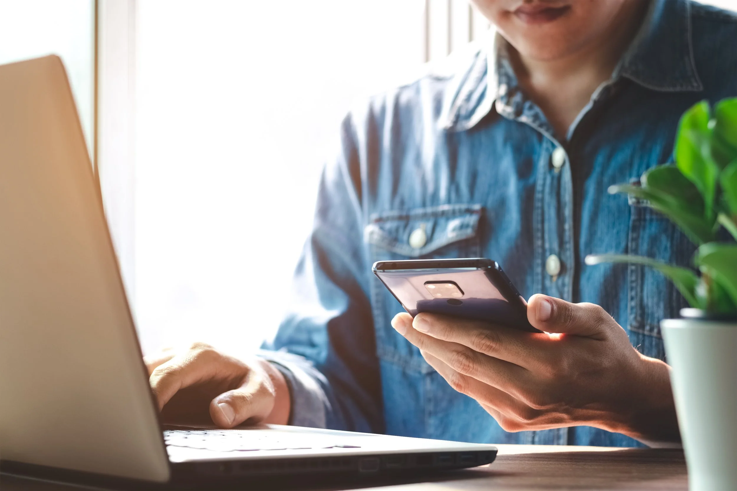 Person wearing a denim shirt looking at a smartphone while sitting at a desk with a laptop and a plant nearby.