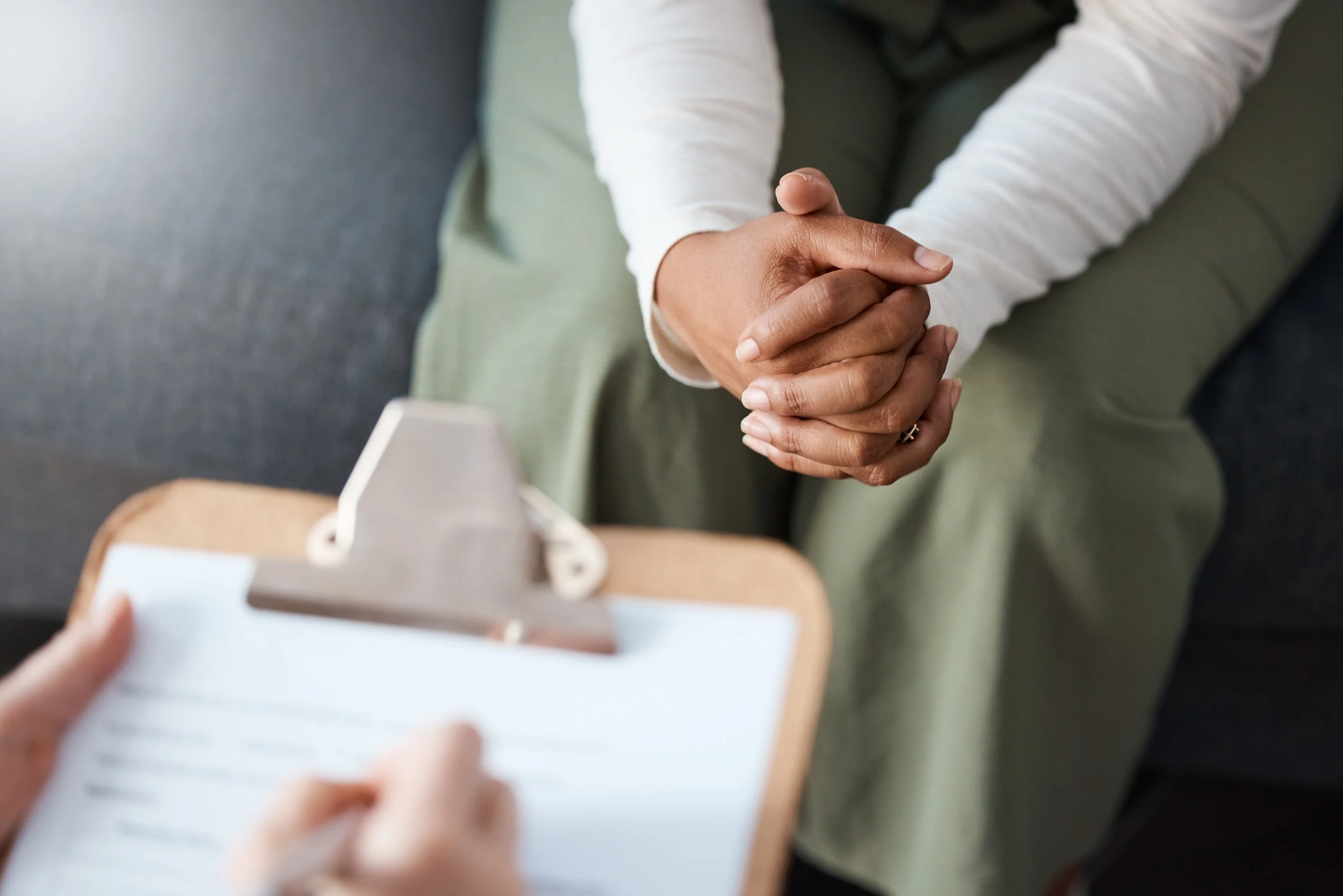 A person with intertwined hands talking to a therapist with a clipboard