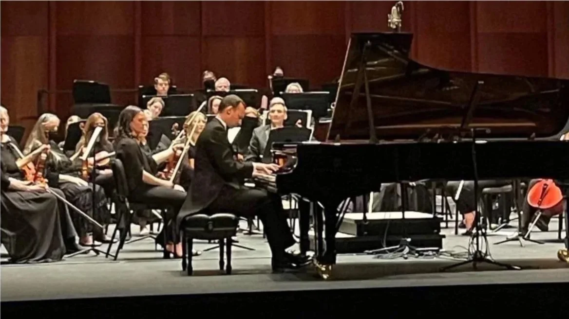 An orchestra performing on stage, with a pianist playing grand piano in front and musicians with various instruments in the background.