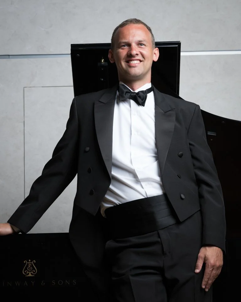A man in a tuxedo smiling and posing next to a grand piano.