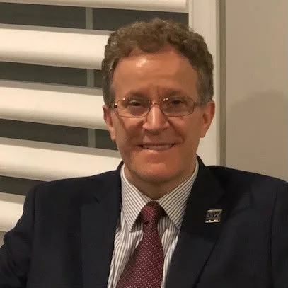 A man with curly hair wearing glasses, a suit, and a tie, smiling at the camera indoors with window blinds in the background.