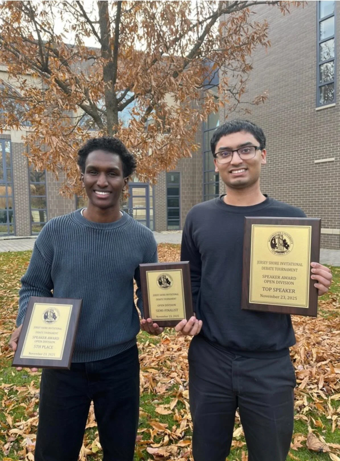 Two young men standing outdoors in autumn with fallen leaves and a building behind them, holding plaques and certificates. The man on the left is smiling and wearing a gray sweater; the man on the right is also smiling and wearing glasses and a black shirt. The plaques indicate they won awards at the Jersey Shore Invitational Debate Tournament.
