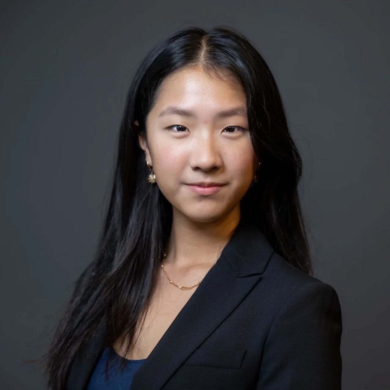 A young woman with long black hair, wearing a black blazer and a blue top, posing against a dark gray background.