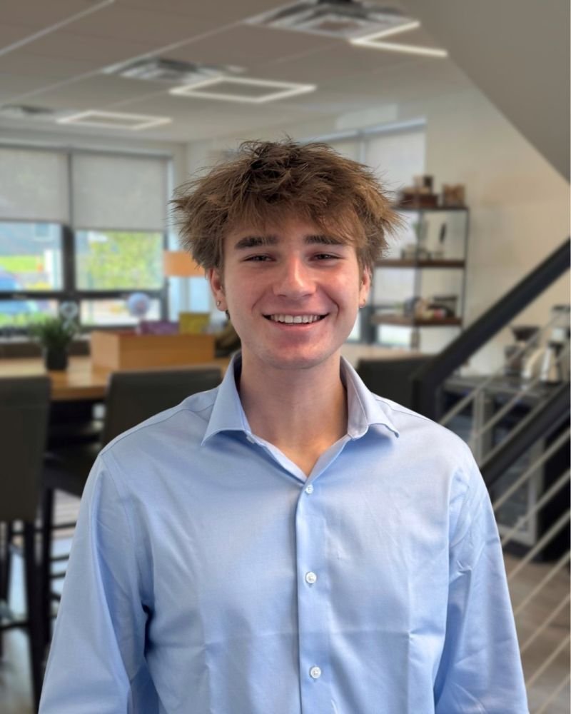 Young man with tousled brown hair and wearing a light blue dress shirt, smiling indoors in a modern office or cafe setting.
