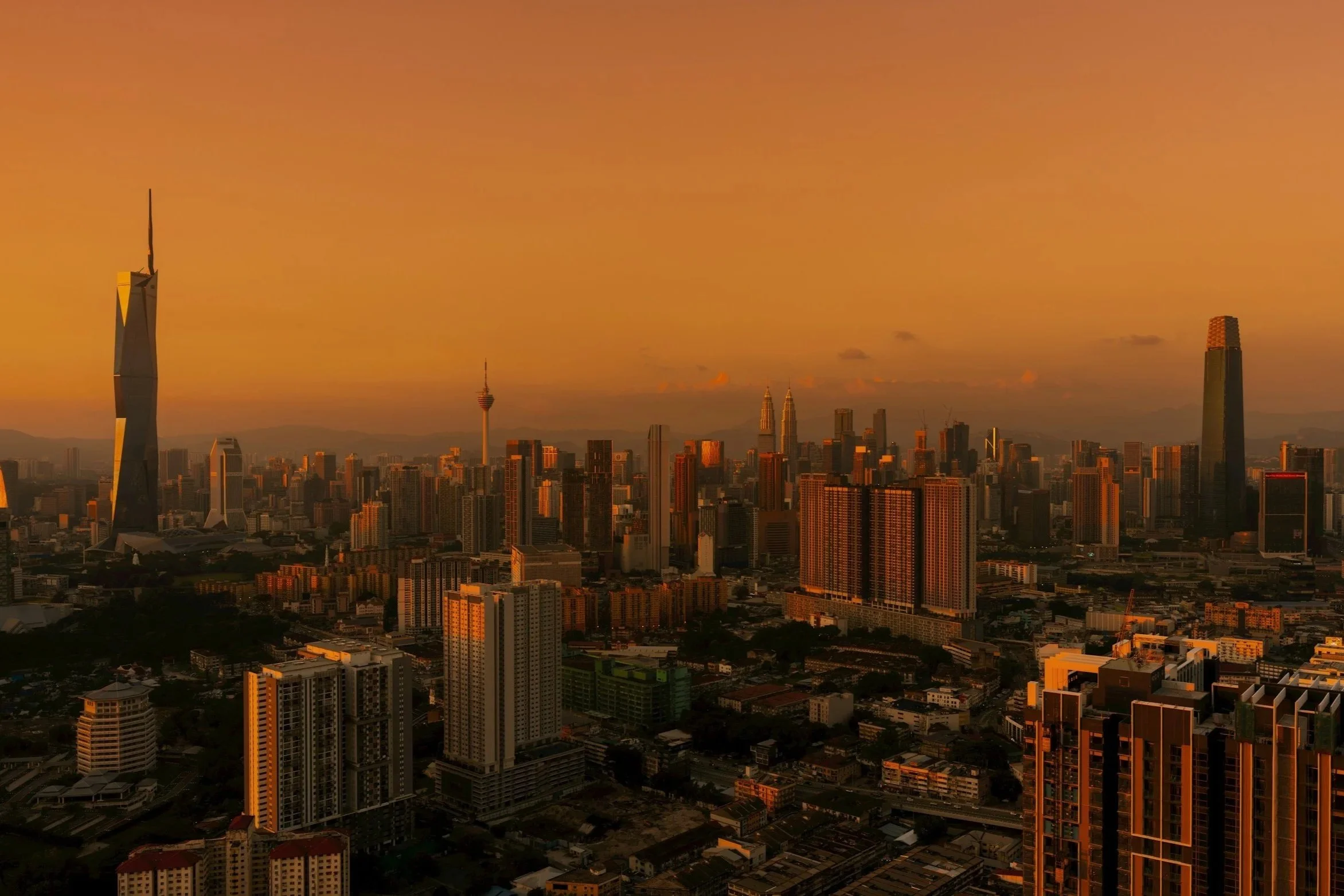 A city skyline at sunset with tall skyscrapers and buildings, including one with a unique modern design, under an orange sky.