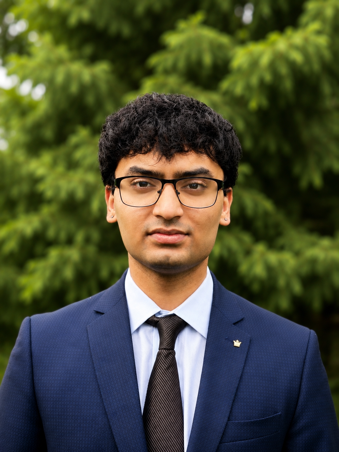 Close-up portrait of a young man with black hair, glasses, wearing a navy blue suit, light blue shirt, and dark tie, standing outdoors in front of green foliage.