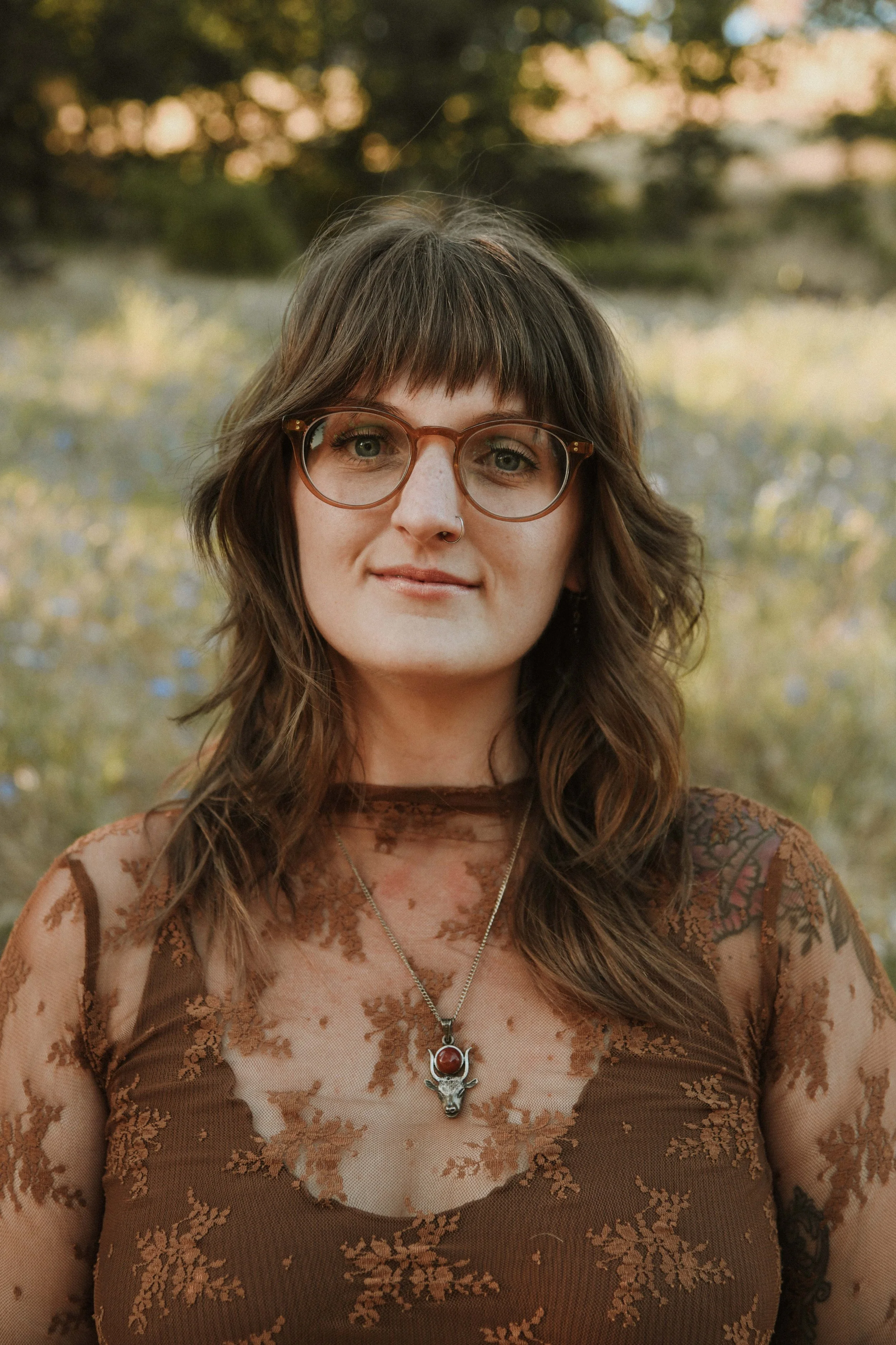 A woman with long wavy brown hair, wearing glasses and a brown lace dress with floral patterns. She has a necklace with a sun or moon pendant and a deer skull with a red stone on her chest. The background features an outdoor setting with blurred trees and soft sunlight.