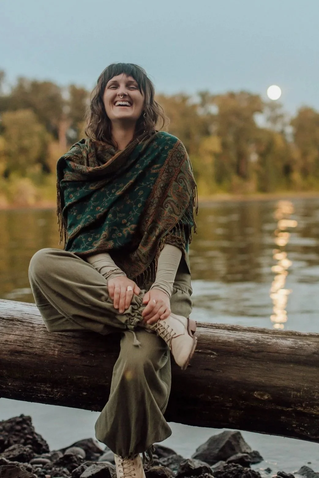 Emily Rose Meredith laughing on a riverside log under a full moon, relational healing and self-trust coach