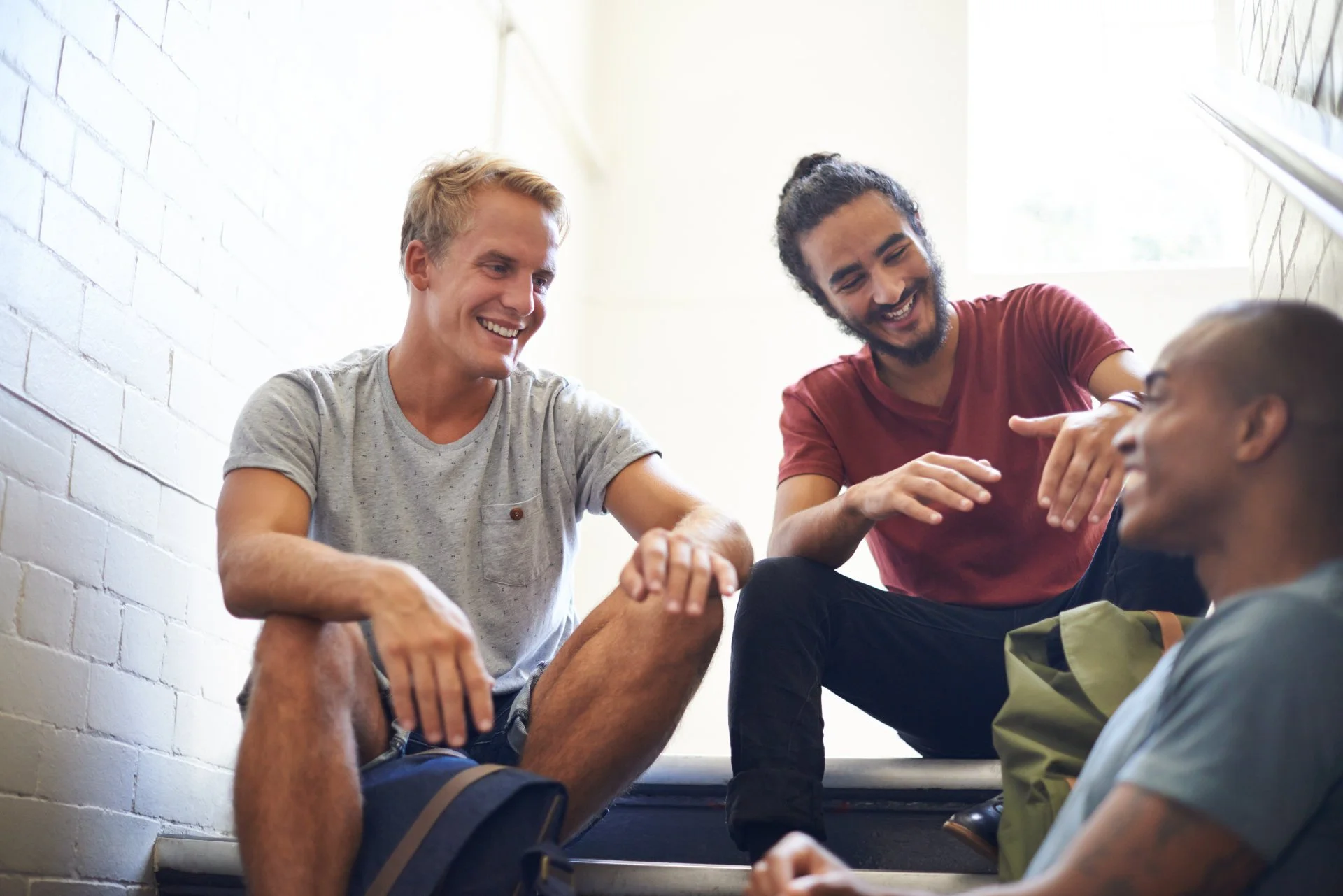 Three young adults sitting and chatting on stairs in a bright room with a white brick wall and a window.