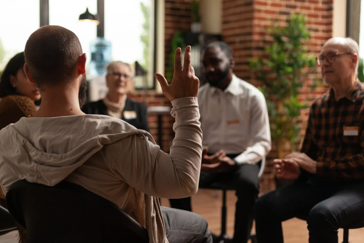 Person raising hand while speaking to a diverse group seated in a circle during a group discussion in a bright room.