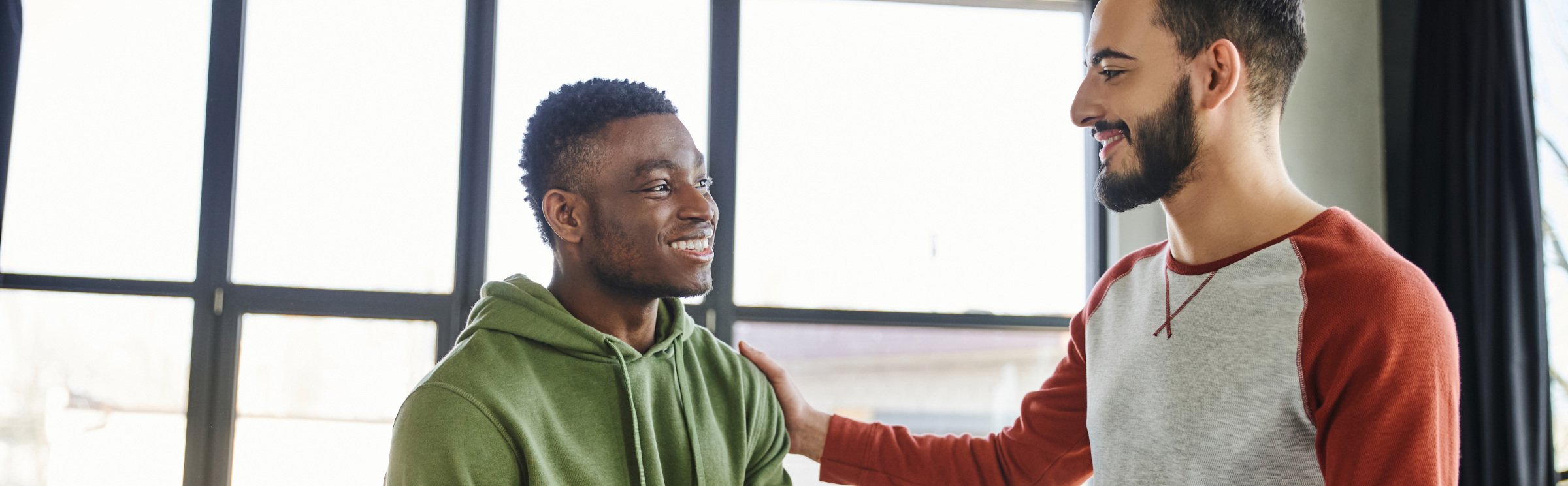 Two men smiling and greeting each other indoors, with large windows in the background.
