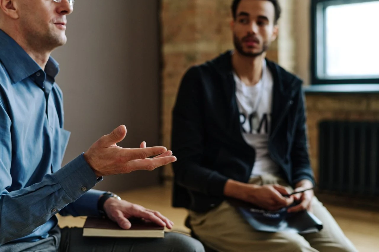 Two men are having a conversation in an indoor setting, with one of them gesturing with his hand while the other holds a smartphone.