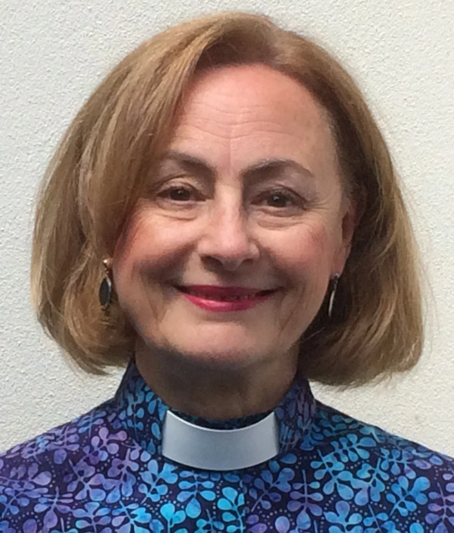 A woman with shoulder-length blond hair, wearing a clerical collar and a blue patterned outfit, standing against a plain white wall.