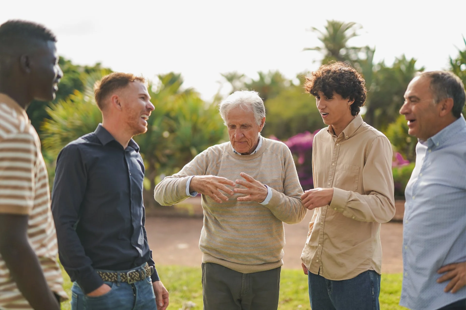 Group of five people, diverse ages and ethnicities, engaged in conversation outdoors with greenery and trees in background