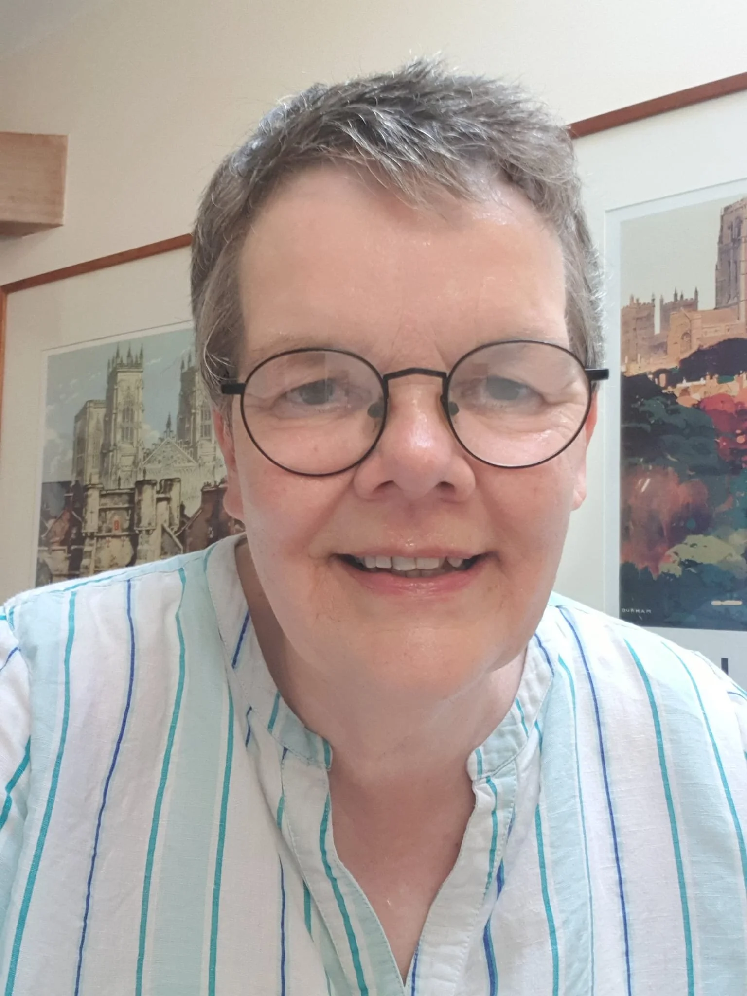 Close-up of a smiling lady with short gray hair and round glasses, wearing a light-colored striped shirt, indoors with artwork of a castle and a historic building in the background.