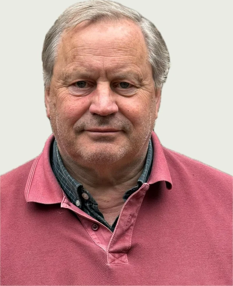 Close-up of a middle-aged man with gray hair and blue eyes, wearing a pink polo shirt over a dark checked collared shirt, standing against a plain light-colored background.