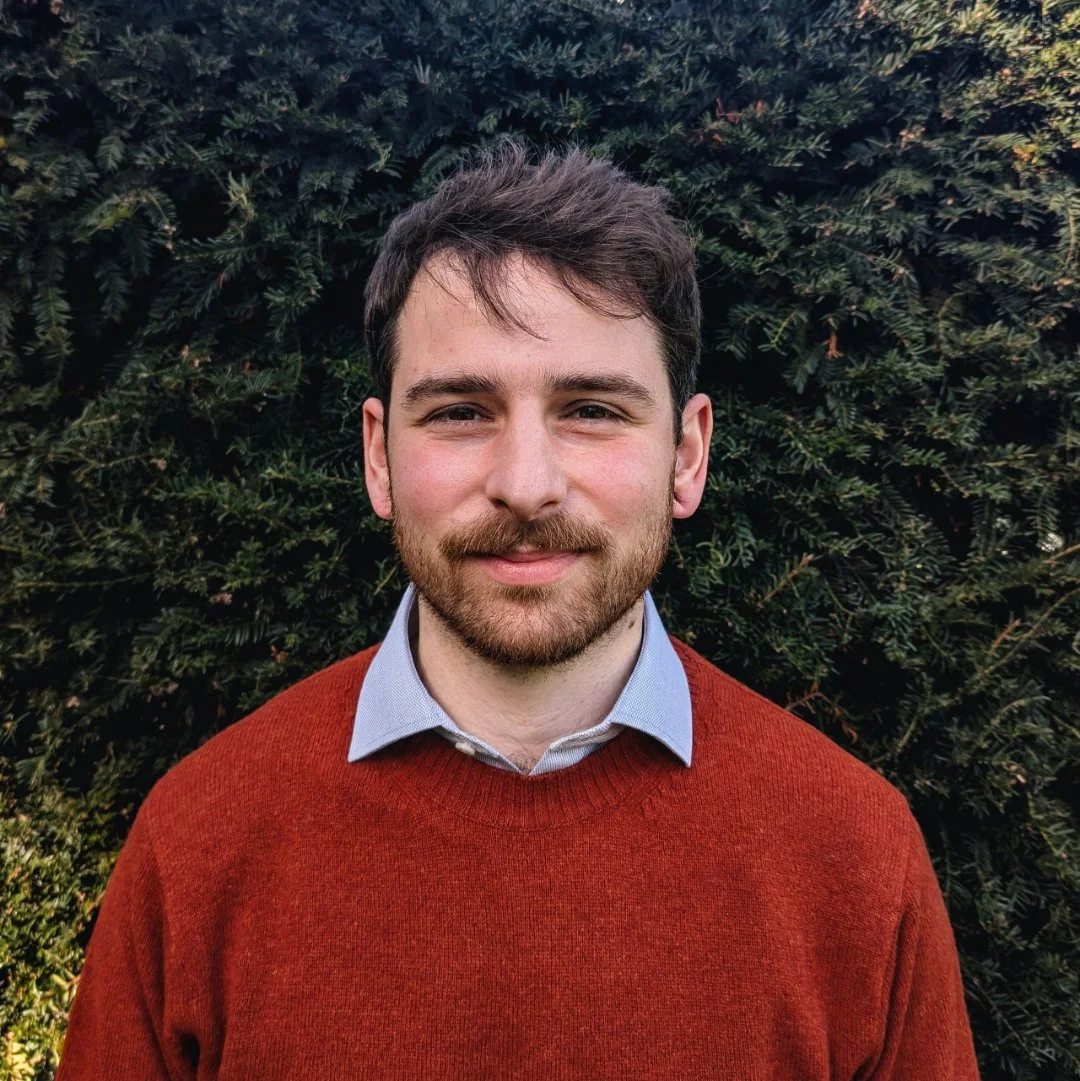 A young man with brown hair and a beard standing outdoors in front of green shrubbery, wearing a red sweater over a collared shirt.