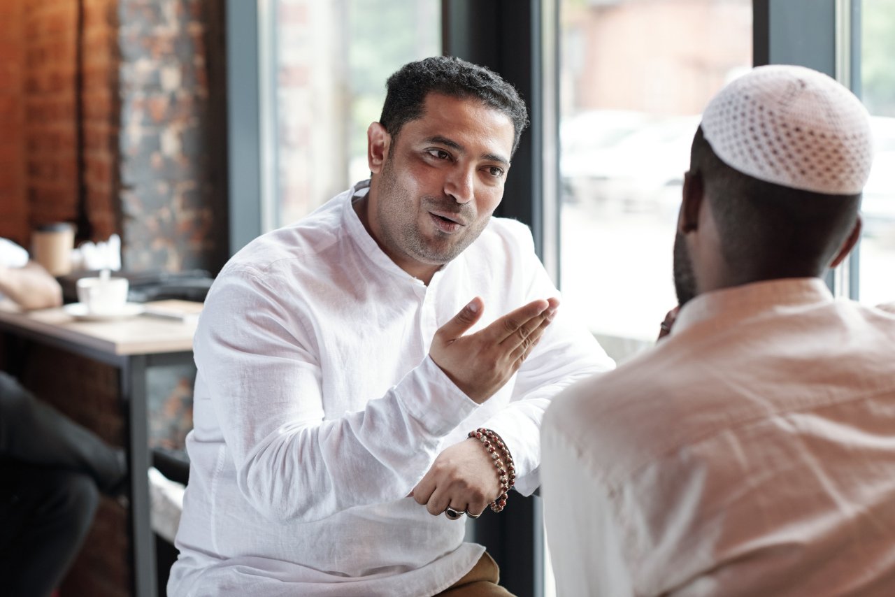 Two men engaged in a conversation, one gesturing with hand, inside a cafe with large windows and exposed brick walls.