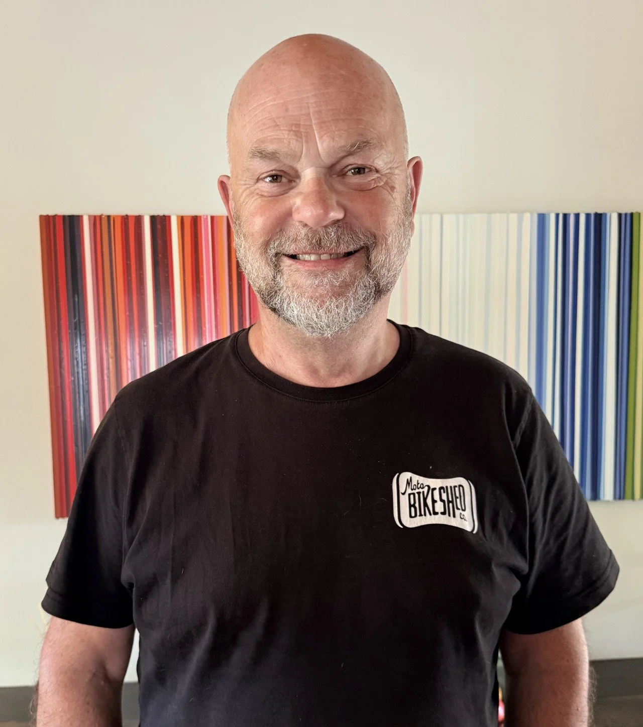 A smiling, middle-aged man with a bald head and beard wearing a black T-shirt with a graphic that reads 'Moto Bikeshed Co.' in front of a colorful, striped artwork.