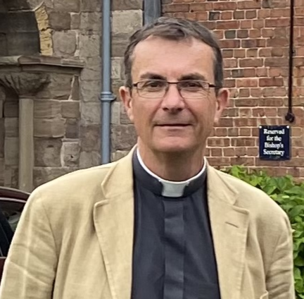A man in clerical clothing, wearing glasses, standing outside in front of a brick and stone building with greenery.