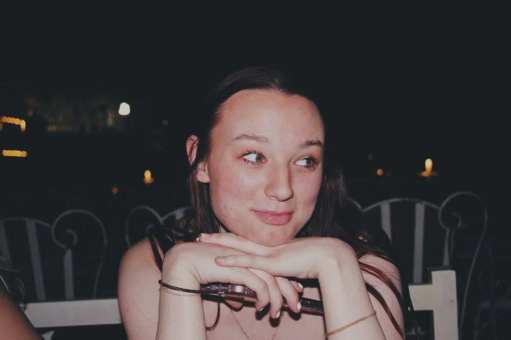 A young woman with brown hair and blue eyes resting her chin on her crossed hands, sitting at an outdoor location at night with outdoor lights in the background.