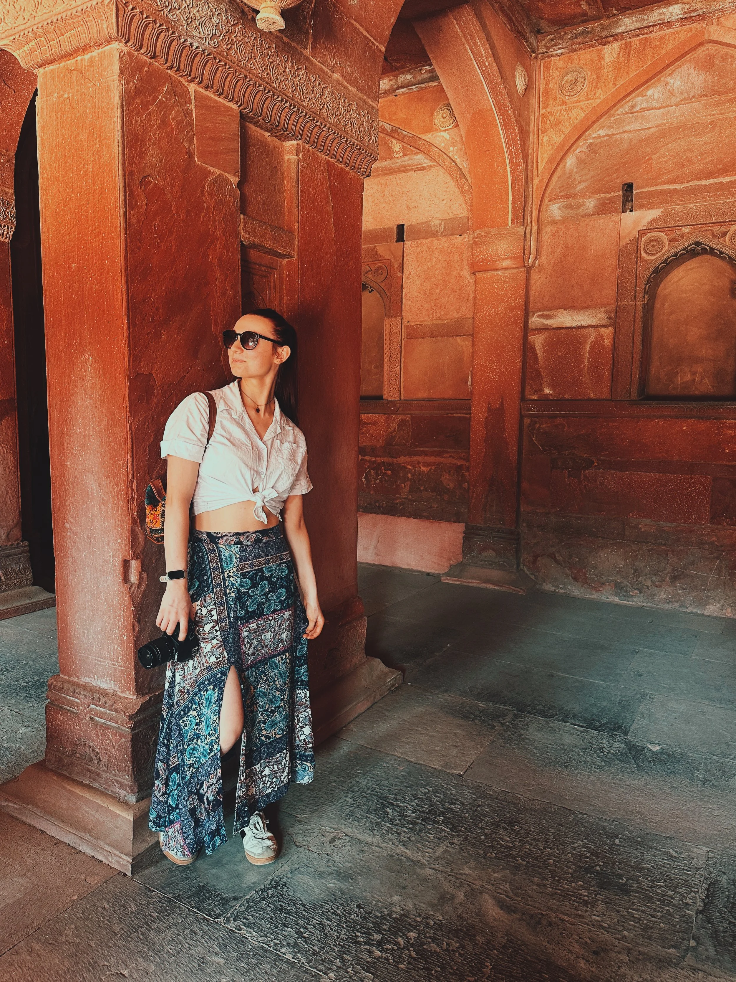 A woman with long dark hair wearing sunglasses, a white tied-up shirt, a long patterned skirt, and sneakers, standing against a reddish-brown stone wall inside an ornate historic building.