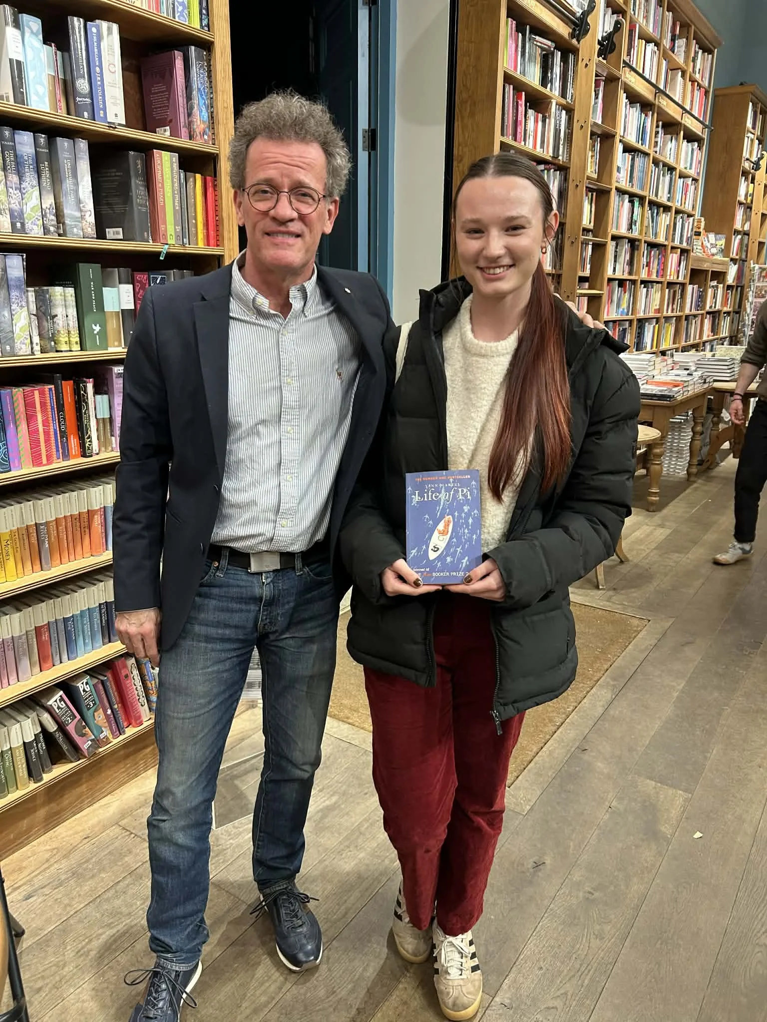 A man with glasses and a woman standing in a bookstore, both smiling at the camera. The woman is holding a blue book titled 'Life of Pi.'