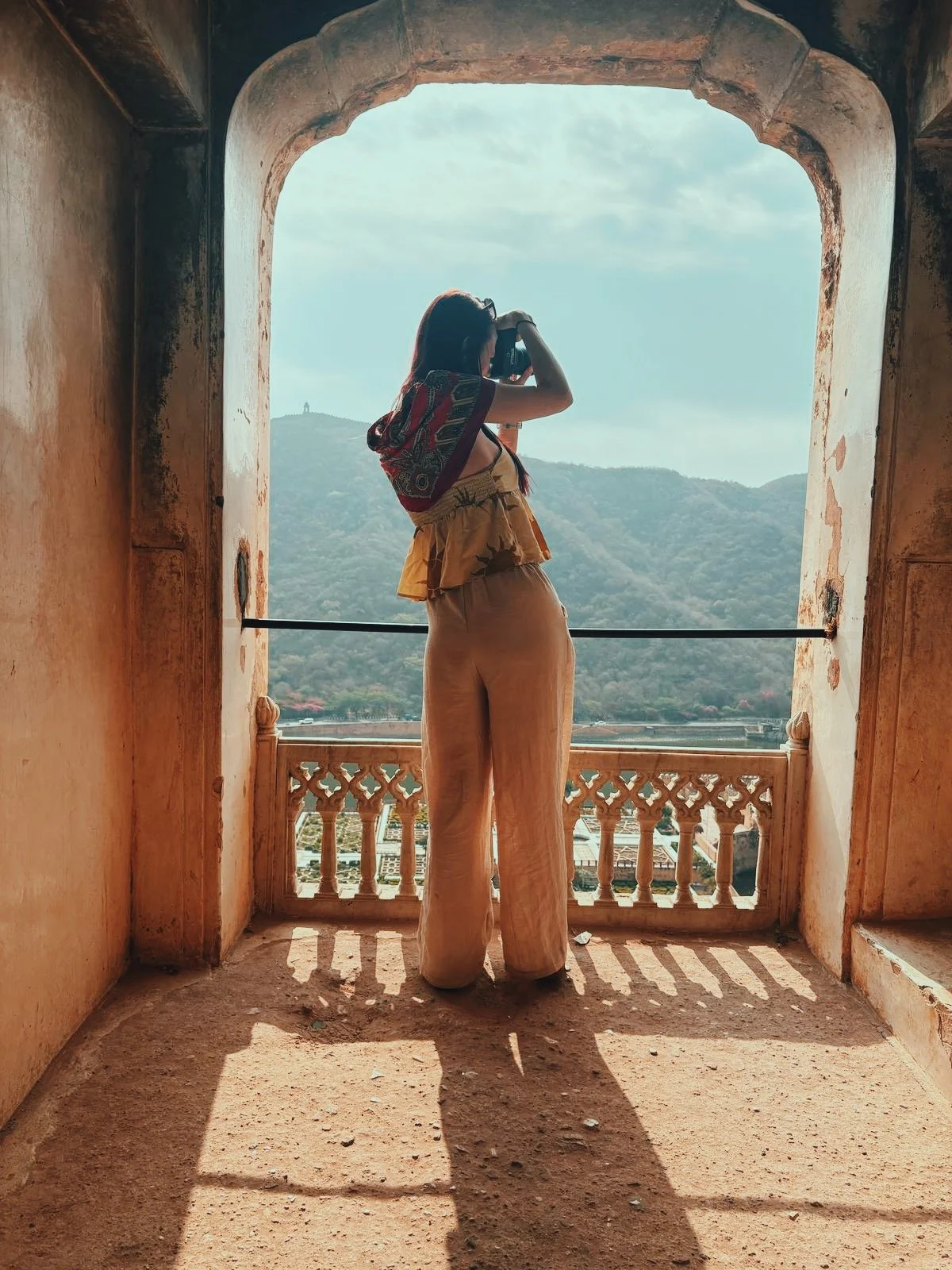 A woman standing on a balcony, taking a photo with a camera, overlooking a mountainous landscape.